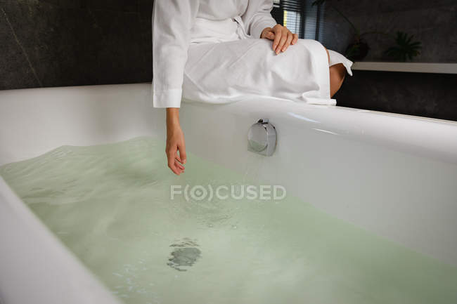 Вид спереди средней части женщины, сидящей на краю ванны и касающейся воды, в современной ванной комнате . — стоковое фото