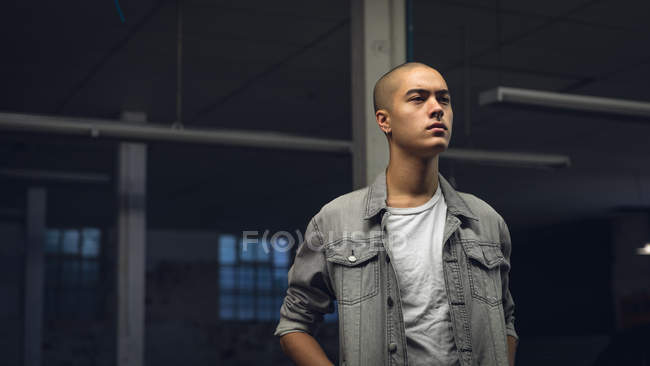 Front view of a young Hispanic-American man with piercings wearing a grey jacket over a white shirt while standing inside a dark and empty warehouse and looking away from the camera — Stock Photo
