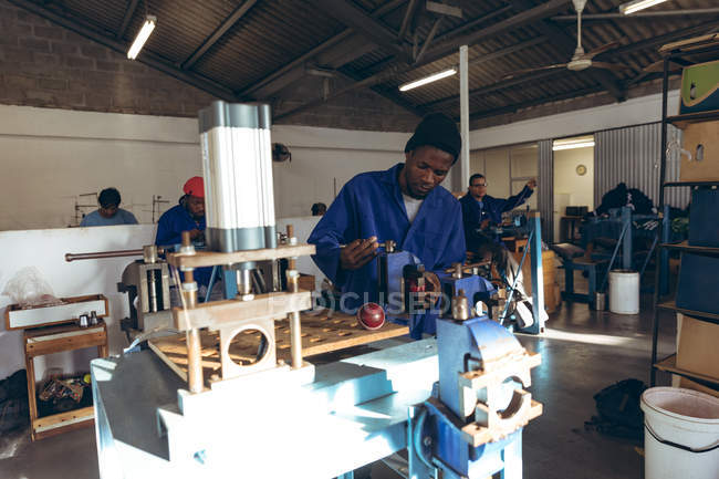 Front view of a young African American man wearing a hat and work clothes operating a machine in a workshop at a factory making cricket balls, in the background colleagues are working on other parts of the production line. — Stock Photo