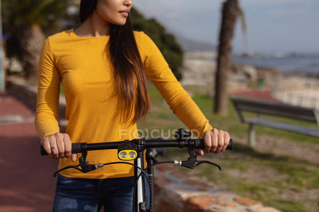 Front view mid section of a young mixed race woman standing outside with her bicycle, a palm tree and a sea view in the background — Stock Photo