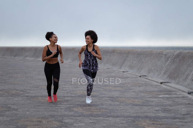 Вид спереду на двох молодих сестер змішаної раси в спортивному одязі, що бігають і дивляться один на одного, посміхаючись у зовнішньому міському просторі — стокове фото