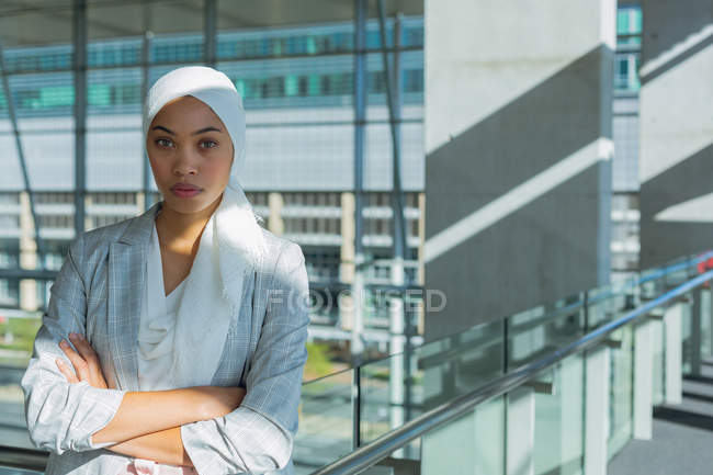 Schöne Geschäftsfrau im Hijab mit verschränkten Armen, die in die Kamera im Flur eines modernen Büros blickt. — Stockfoto