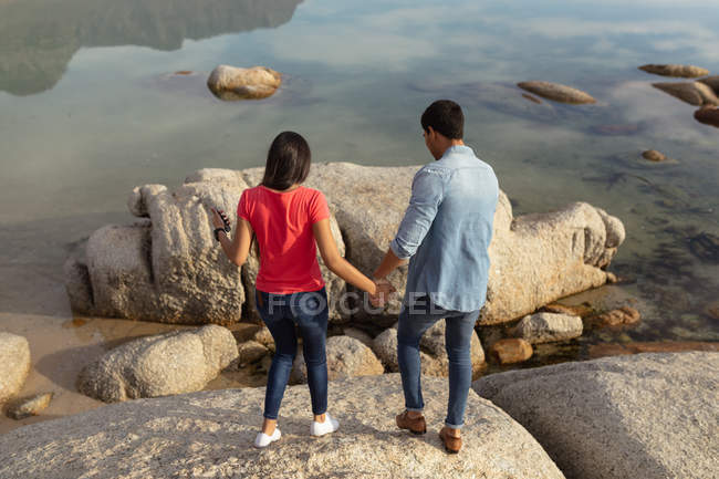 Vista posteriore elevata di una giovane coppia mista che si tiene per mano in piedi su rocce in riva al mare — Foto stock