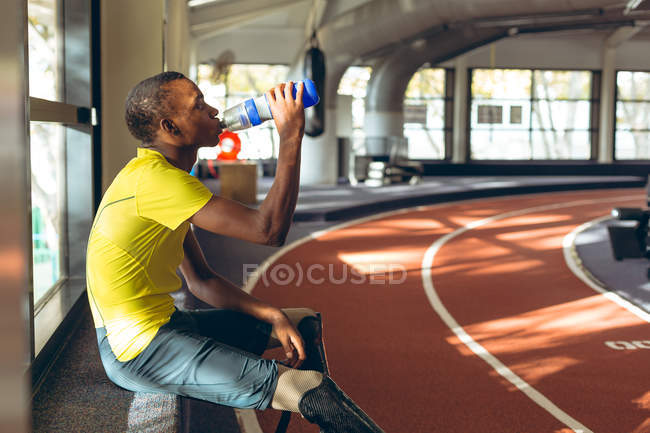 Vue latérale de l'eau potable sportive masculine afro-américaine handicapée sur une piste de course dans un centre de remise en forme — Photo de stock