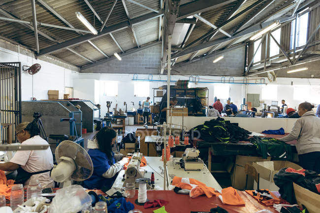 Side view of a diverse group of female colleagues working at sewing machines and sorting through fabric at a sports clothing factory, with other colleagues working in the background. — Stock Photo