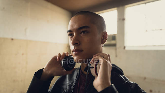 Side view of a young Hispanic-American man wearing a black leather jacket while holding headphones inside an empty warehouse — Stock Photo