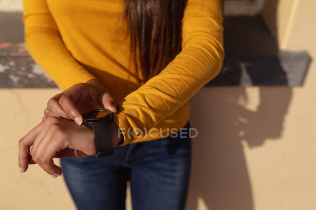 Visão frontal seção média da mulher usando seu smartwatch encostado a uma parede do lado de fora ao sol — Fotografia de Stock
