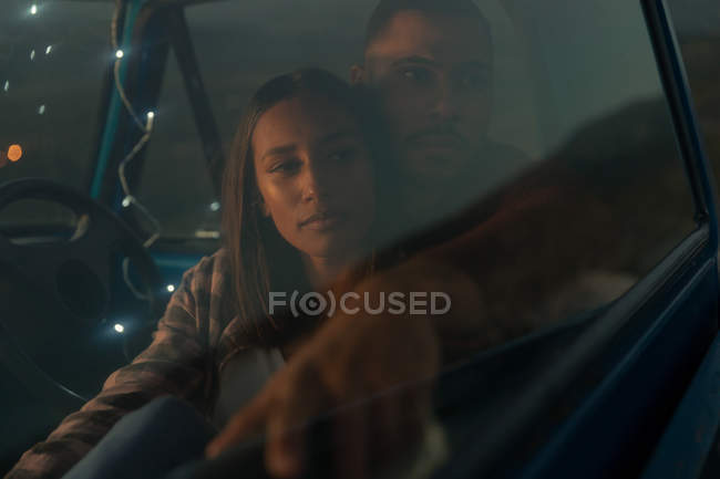 Close up front view of a young mixed race couple sitting in their pick-up truck, smiling and embracing at dusk during a stop off on a road trip. — Stock Photo