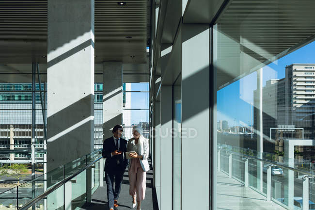Vista frontal de empresários que trabalham em tablet digital enquanto caminham no corredor no escritório moderno . — Fotografia de Stock