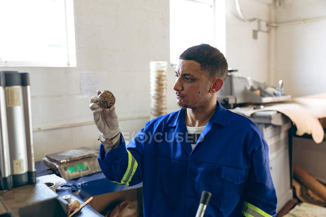 Vue de face rapprochée d'un jeune homme de race mixte portant des gants et des combinaisons tenant le cœur d'une balle et la vérifiant dans une usine fabriquant des balles de cricket, avec des équipements et des matériaux visibles en arrière-plan — Photo de stock