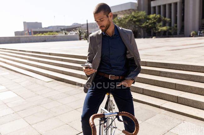 Vista frontal de cerca de un joven caucásico usando un teléfono inteligente, sentado en su bicicleta en una calle de la ciudad. Nómada digital en movimiento . - foto de stock