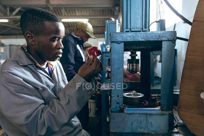 Vista lateral de cerca de un joven trabajador afroamericano sentado, sosteniendo cuero rojo en forma y operando una máquina en una fábrica haciendo pelotas de cricket, en el fondo se puede ver a un colega trabajando junto a él en la línea de producción . - foto de stock