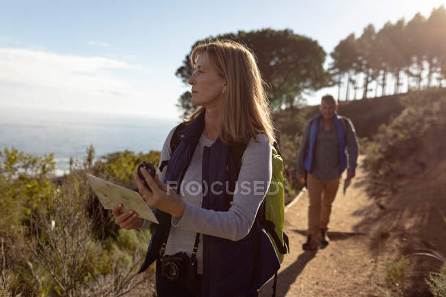 Front view close up of a mature Caucasian woman holding a map and a compass looking away to get her bearings and man wearing backpacks walking single along a trail during a hike. — Stock Photo