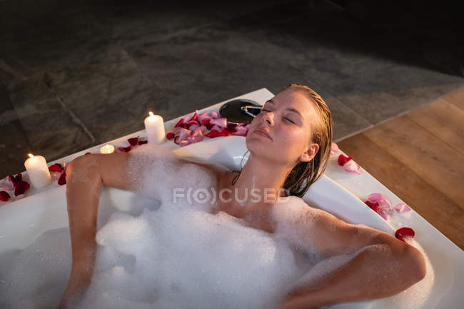 Elevado primer plano de una joven mujer caucásica acostada en un baño de espuma con velas encendidas y pétalos de rosa a su alrededor . - foto de stock