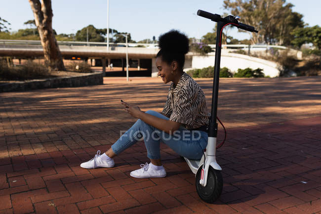 Vue latérale d'une jeune femme souriante de race mixte assise sur un scooter électrique dans un parc urbain à l'aide d'un smartphone — Photo de stock
