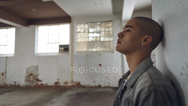 Seitenansicht eines jungen hispanisch-amerikanischen Mannes mit Piercings, der eine graue Jacke trägt, während er sich an eine weiße Wand in einer leeren Lagerhalle lehnt und von der Kamera wegschaut — Stockfoto