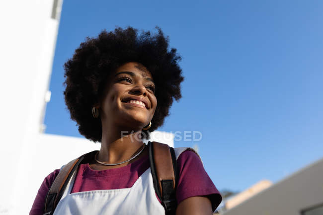 Baixo ângulo fechar-se de uma jovem mulher de raça mista de pé e olhando para longe sorrindo contra o céu azul — Fotografia de Stock