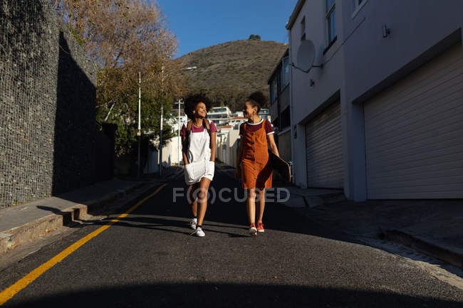 Vista frontal de dos hermanas adultas jóvenes de raza mixta, una con una mochila y la otra con un monopatín, hablando y sonriendo mientras caminan en una calle bajo el sol - foto de stock