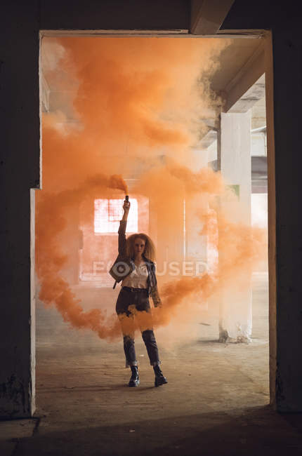 Vista frontale di una giovane donna caucasica con i capelli ricci che indossa una giacca di pelle mentre guarda attentamente la fotocamera e tiene in mano un fumatore che produce un fumo arancione all'interno di un magazzino vuoto — Foto stock