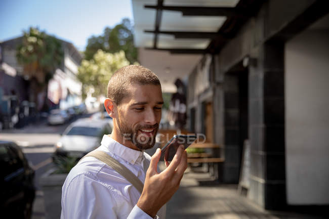 Vista laterale da vicino di un giovane caucasico sorridente che parla su uno smartphone tenendolo davanti al viso, in piedi in una strada della città. Nomade digitale in movimento . — Foto stock