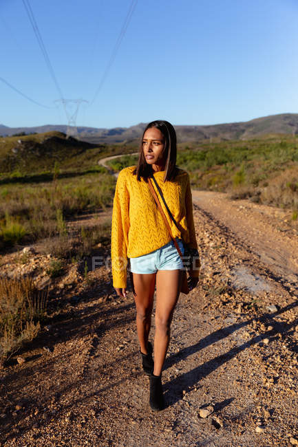 Vue de face d'une jeune femme métissée marchant le long d'un sentier à travers un paysage rural ensoleillé, avec des montagnes à l'horizon en arrière-plan. Elle porte un short, avec un haut jaune et un sac à main . — Photo de stock