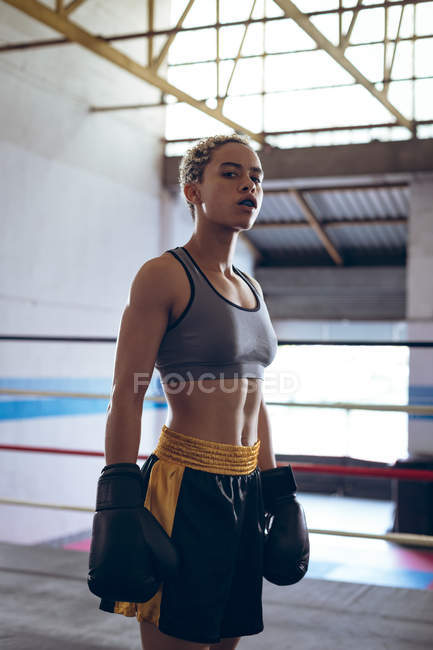 Vista lateral do boxeador feminino com luvas de boxe em pé no ringue de boxe no clube de boxe. Forte lutador feminino no treinamento de ginásio de boxe duro . — Fotografia de Stock