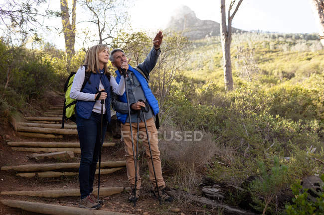 Front view of a mature Caucasian woman and man wearing backpacks and using Nordic walking sticks, stopping on a downhill trail to admire the view and pointing to the distance during a hike — Stock Photo