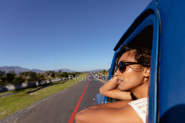 Close up side view of a young mixed race woman wearing sunglasses leaning out of the front passenger side window of a pick-up truck as it drives down the highway on a road trip — Stock Photo