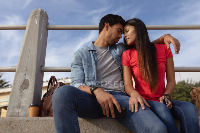 Vue de face gros plan d'un jeune couple mixte assis à l'extérieur sur un mur embrassant les yeux fermés — Photo de stock