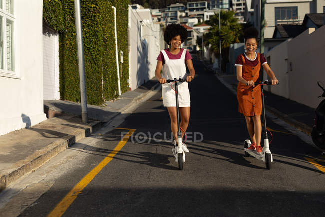Front view of two young adult mixed race sisters smiling and riding scooters in a street in the sun — Stock Photo