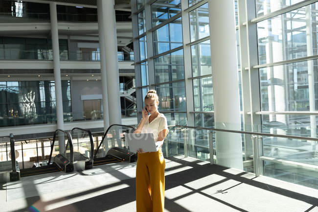Vista frontale della giovane donna d'affari caucasica in piedi in una moderna lobby aziendale che tiene il computer portatile e parla su smartphone . — Foto stock