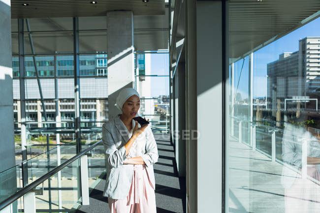 Vista frontal de la mujer de negocios en hijab mirando a través de la ventana mientras habla por teléfono móvil en el pasillo en la oficina
. - foto de stock
