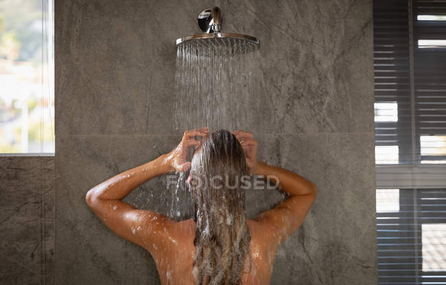 Вид сзади на молодую кавказскую женщину, стоящую под душем в современной ванной . — стоковое фото