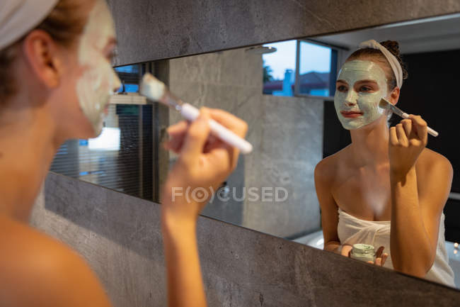 Над плечем зору молода кавказька жінка дивиться на дзеркало і застосовуючи маску для обличчя щіткою у сучасній ванній. — стокове фото