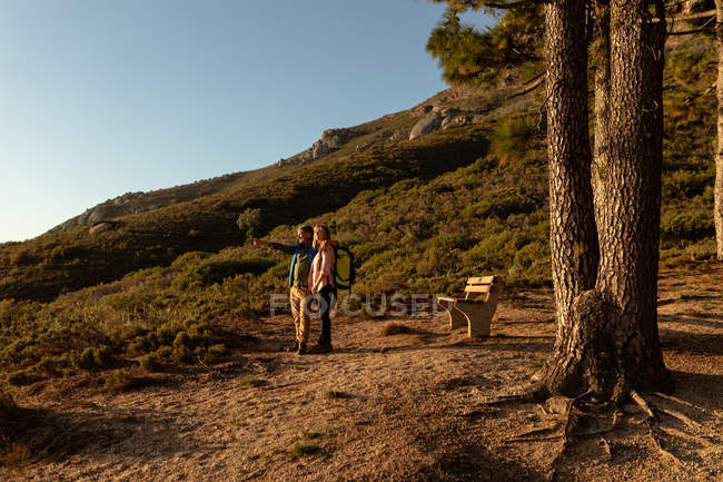 Vista lateral de um homem e uma mulher caucasianos maduros usando mochilas parando para apreciar o cenário e apontando para a distância durante uma caminhada
. — Fotografia de Stock