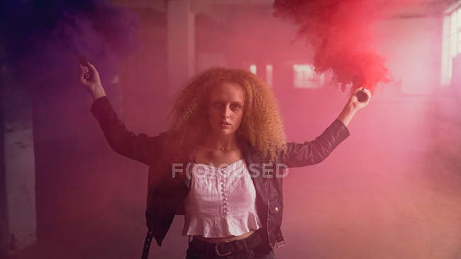 Front view of a young Caucasian woman with curly hair wearing leather jacket while looking intently at the camera and holding a smoke maker producing red and blue smoke inside an empty warehouse — Stock Photo