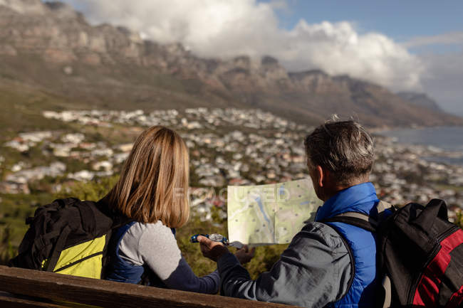 Rear view close up of a mature Caucasian woman and man wearing backpacks sitting on a bench and looking at a map and a compass which the man is holding during a hike — Stock Photo