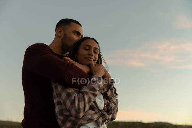 Side view of a young mixed race couple sitting outside embracing at dusk during a stop off on a road trip. The woman is smiling and has her eyes closed. — Stock Photo