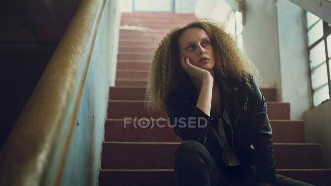 Vue à angle bas d'une jeune femme caucasienne aux cheveux bouclés portant une veste en cuir assise dans les escaliers avec la main sur le menton — Photo de stock