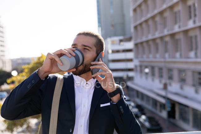 Vue de face gros plan d'un jeune homme caucasien buvant un café à emporter et parlant sur son smartphone, debout dans la ville. Nomade numérique en mouvement . — Photo de stock