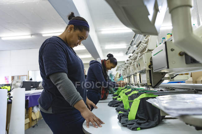 Side view of two young mixed race women checking automated sewing machines stitching shirts in a sports clothing factory. — Stock Photo