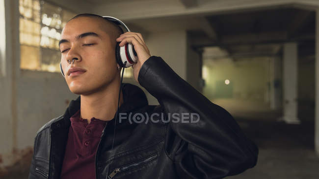 Side view of a young Hispanic-American man wearing a black leather jacket over a maroon shirt with hand on headphones and closed eyes while standing inside an empty warehouse — Stock Photo