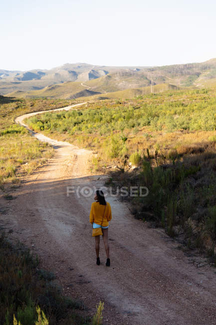 Visão traseira de uma jovem mista caminhando ao longo de uma trilha através de uma paisagem rural ensolarada em direção a montanhas no horizonte . — Fotografia de Stock