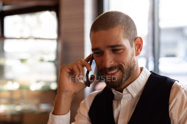Vista frontal perto de um jovem caucasiano sorridente em um telefonema sentado em uma mesa dentro de um café. Digital Nomad em movimento . — Fotografia de Stock