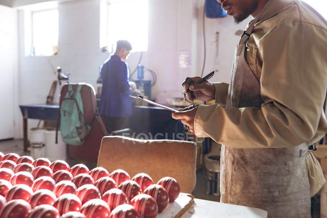 Visão lateral no meio da seção de um jovem afro-americano segurando uma prancheta e escrevendo enquanto ele verifica fileiras de bolas de críquete no final da linha de produção em uma fábrica de equipamentos esportivos. Em segundo plano, um colega trabalha na linha de produção . — Fotografia de Stock