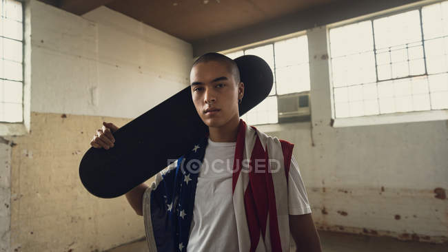 Vista frontale di un giovane ispano-americano che indossa una camicia bianca con uno skateboard nero sulle spalle e una bandiera americana che guarda attentamente la telecamera all'interno di un magazzino vuoto — Foto stock