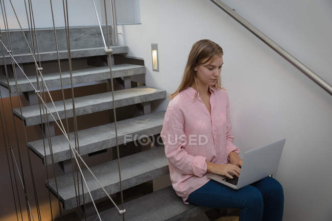 Vista lateral de una joven mujer caucásica usando una camisa rosa, sentada en una escalera en un apartamento usando una computadora portátil . - foto de stock