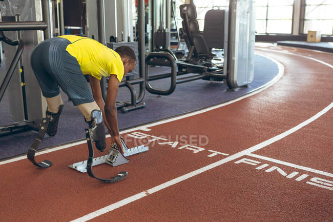 Vista lateral do atlético masculino afro-americano com deficiência no início do bloco na pista de corrida no centro de fitness — Fotografia de Stock