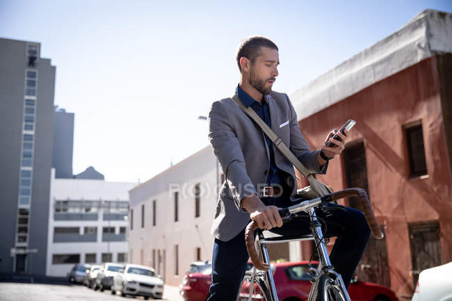 Vista frontal de cerca de un joven caucásico usando un teléfono inteligente, sentado en su bicicleta en una calle de la ciudad. Nómada digital en movimiento . - foto de stock