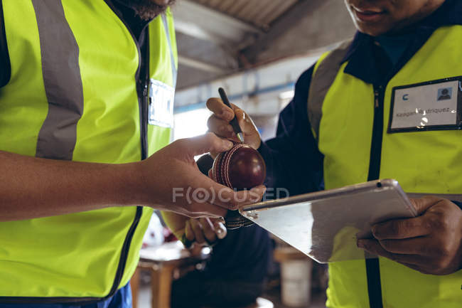 Sección media de un joven mánager masculino de raza mixta sosteniendo una tableta hablando con un trabajador masculino de raza mixta e inspeccionando una pelota que sostiene, en una fábrica de bolas de cricket . - foto de stock